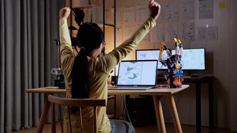 vista trasera de una adolescente asiática con impresión 3d celebrando el éxito en el diseño de una mano cyborg en una computadora portátil en casa