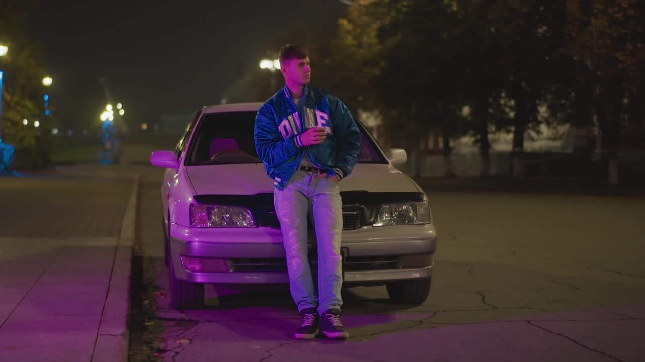 hombre blanco joven apoyado en un coche bajo una luz púrpura de neón, calle urbana vacía de noche, con chaqueta y vaqueros de denim, ambiente reflexivo, faros brillantes, farolas y árboles a lo lejos, cinematográfico