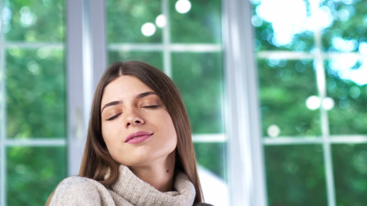 Attractive young lady sitting with her eyes closed. Calm relaxed girl in warm sweater near the window with green trees in it. Blurred backdrop.