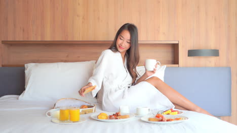 beautiful asian girl in bathrobe drinking coffee with toast and having breakfast in bed in hotel room, plates with fruits and juices on a made-up bed, slow-motion