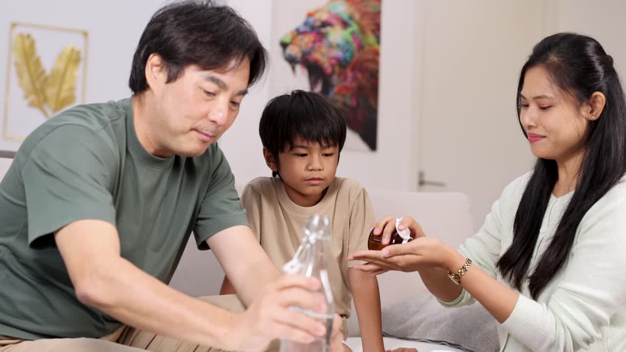 Asian parents help their young son take medicine in a bright, caring living room setting