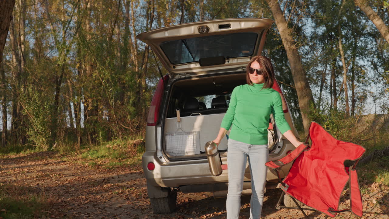 Donna caucasica versa caffè dal thermos. Maglione verde e occhiali da sole, tiene una sedia pieghevole rossa, si allontana dal portellone aperto, luce solare autunnale, routine mattutina del campeggio, energica e sola