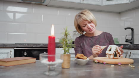 mujer cubriendo cuidadosamente la galleta de pan de jengibre mientras limpia el exceso de hielo de su mano, atmósfera festiva con vela roja, planta en olla y cuenco de galletas