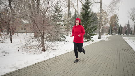 athlete in red beanie and hoodie jogging outdoors during winter on serene park pathway surrounded by snow-covered trees, evergreen pines, and a foggy atmosphere, showcasing endurance
