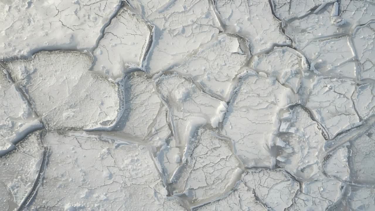 Panoramica aerea iniziale, la camera inquadra le placche di fango secco e screpolato nella distesa fangosa, rivelando microfratture