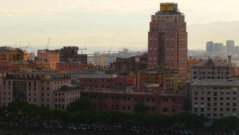 drone shot facing colorful genoa buildings with parked cars below and the port visible in the background at sunset
