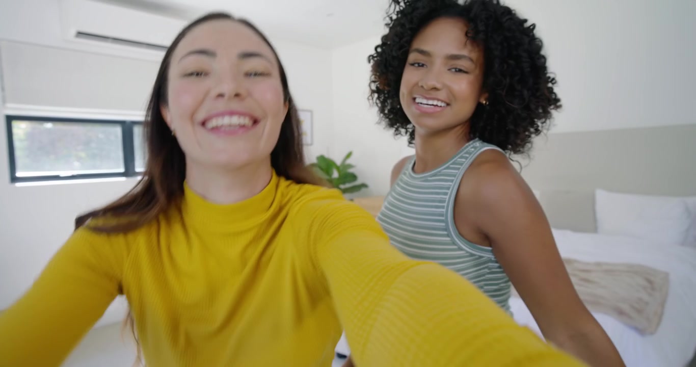 Amiche di diverse etnie che tengono il telefono, inquadrando per un selfie in camera da letto, con una maglietta gialla e una canotta verde.