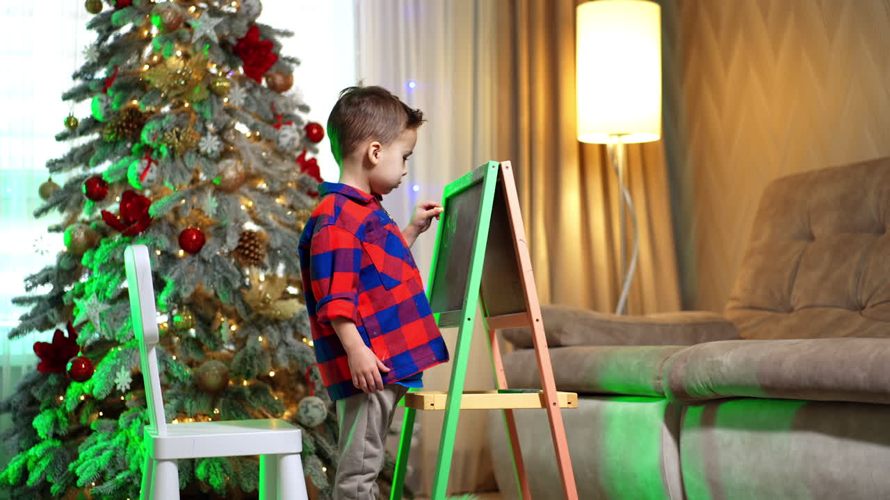 Toddler boy in checkered shirt stands writing on the blackboard. Beautiful Christmas tree at backdrop