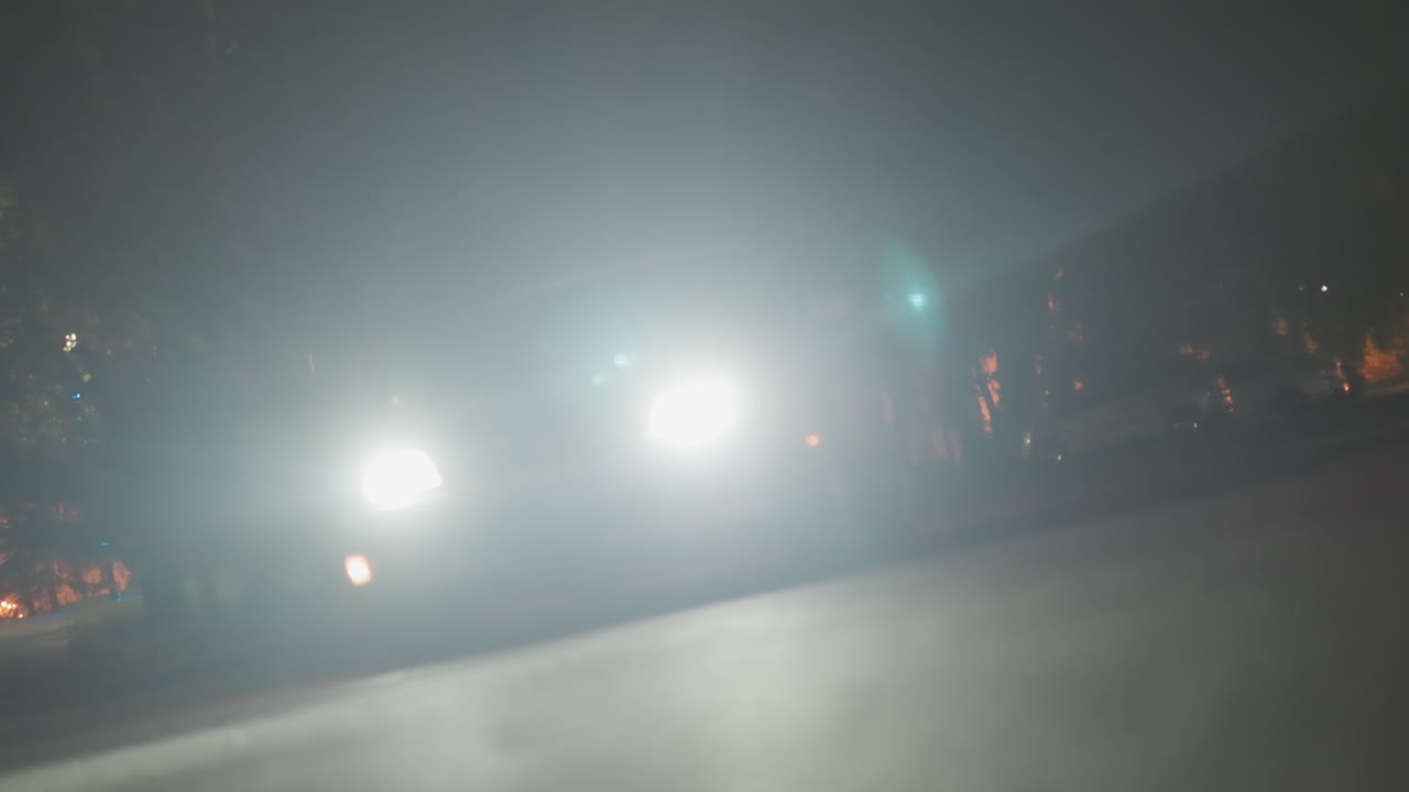 텅 빈 공원 길을 질주하는 밝은 헤드라이트, 강렬한 눈부심과 렌즈 플레어가 있는 로우 앵글 뷰, 도로를 따라 늘어선 나무와 멀리 보이는 건물 조명, 추격과 긴박함, 젖은 아스팔트 바닥에 반사되는 빛