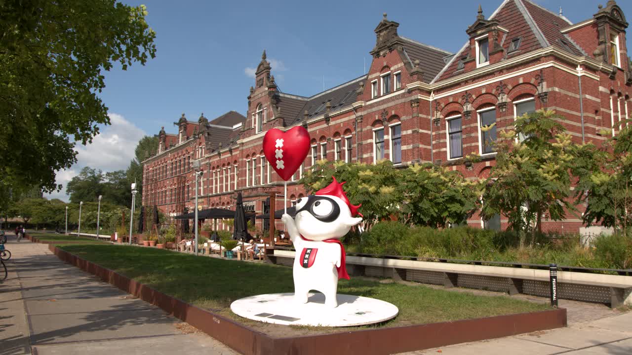 Cartoon mascot with red balloon stands in sunny plaza before ornate brick building, camera pans