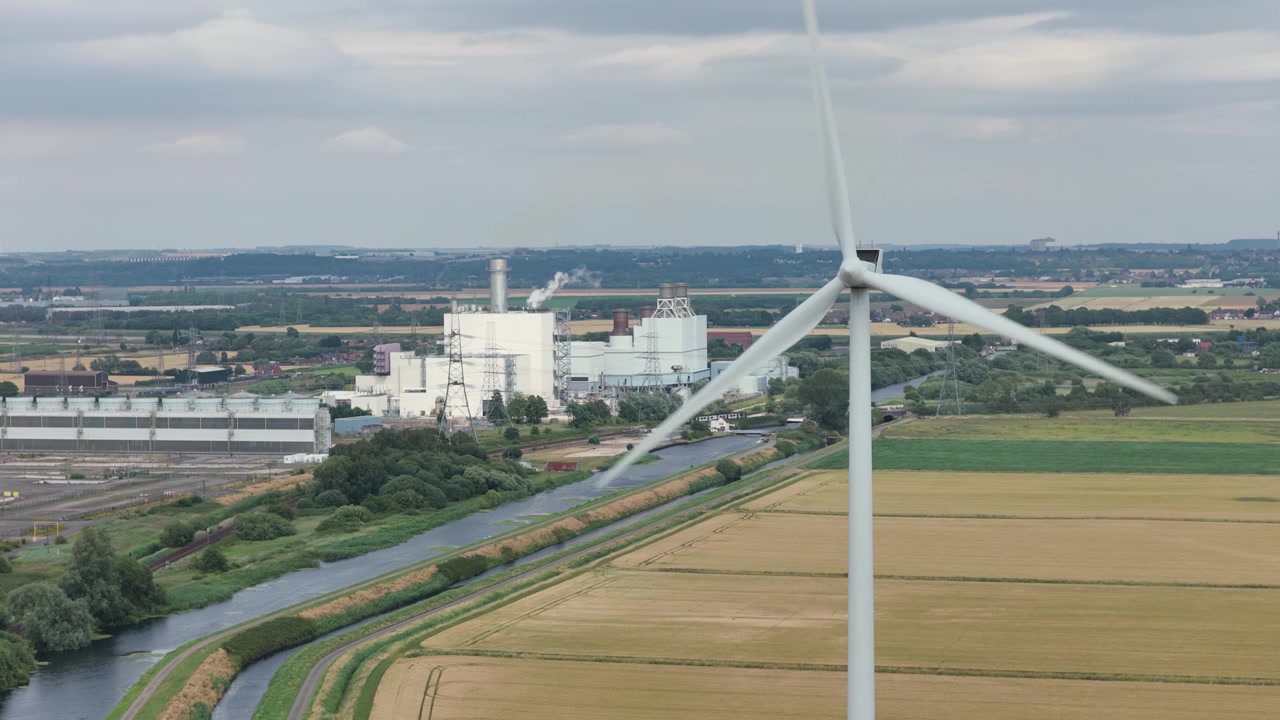 Powerful wind turbine spins, generating clean energy. Industrial buildings border a winding canal and golden agricultural fields under a grey, cloudy daytime sky.
