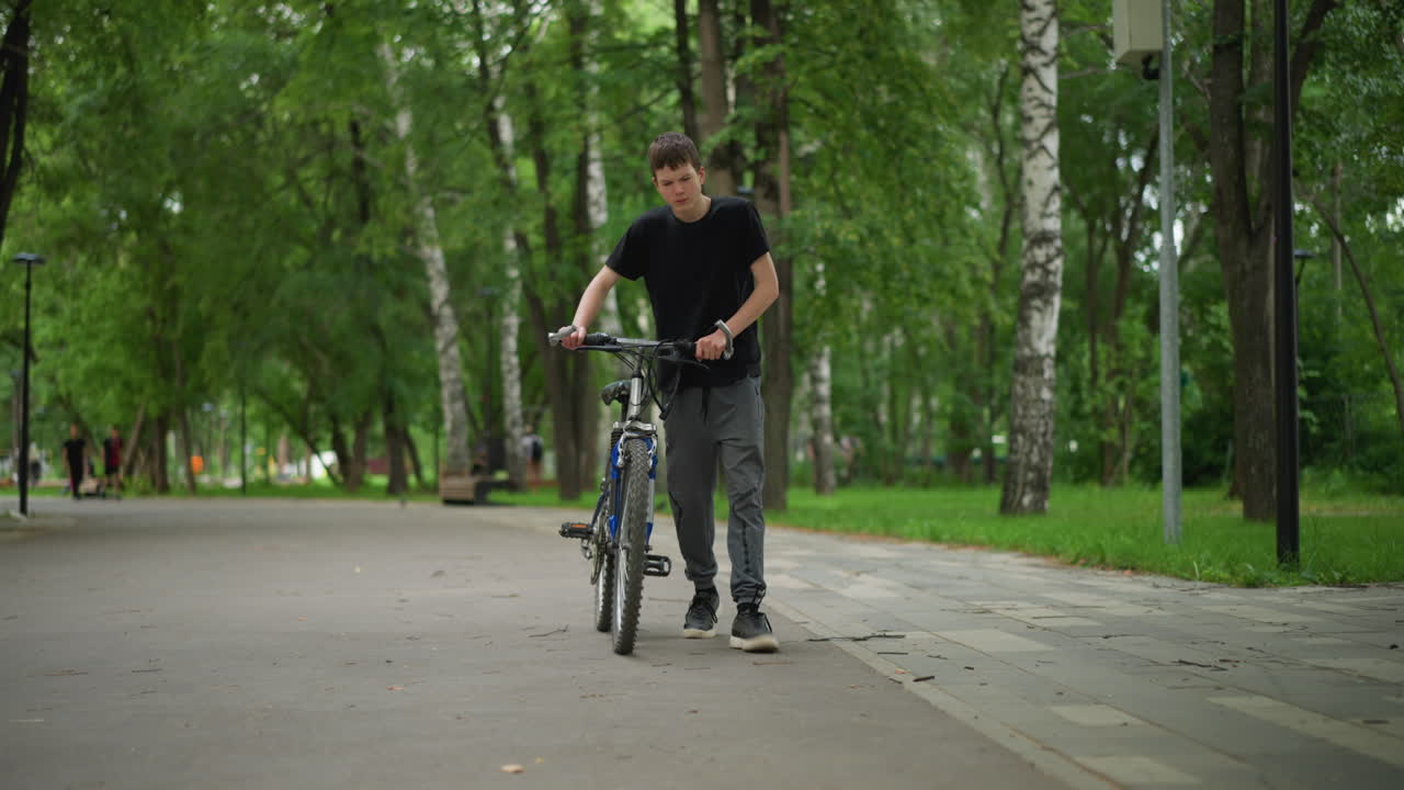 검은 옷을 입은 젊은 백인 소년은 푸른 나무와 자연으로 둘러싸인 평화로운 공원 길을 따라 자전거를 고 있습니다.