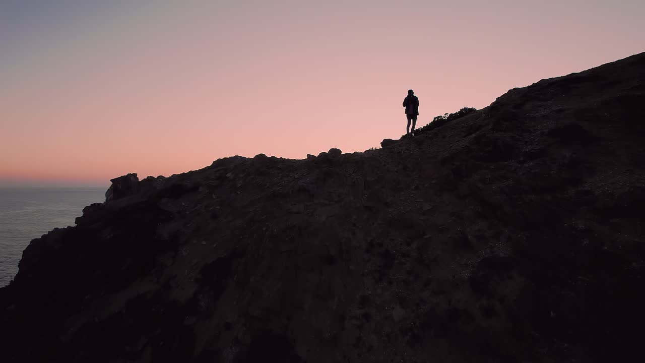 Silhouette of man standing on high coastal cliff in Costa Vicentina Natural Park near São Teotónio Odemira Alentejo Portugal as drone pushes forward revealing Atlantic Ocean horizon at sunset