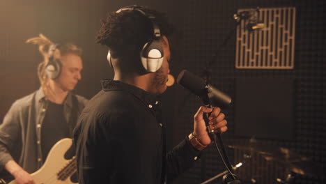 african american vocalist sings lyrical composition into microphone in soundproof room. cool musical band records song in sound recording studio. man plays on guitar at background. music production.