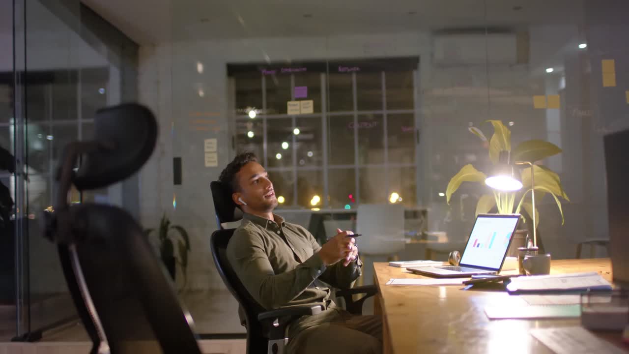 Stanco ma felice uomo d'affari birazziale che ascolta musica con gli auricolari in ufficio di notte, al rallentatore.