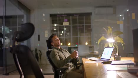 tired happy biracial businessman listening to music with earphones in office at night, slow motion