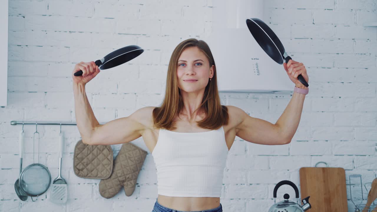 Pretty female with frying pans at home. Muscular young woman holding two empty frying pans. Healthy housewife posing to camera.