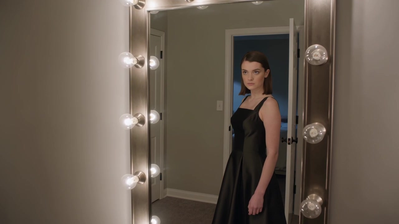 Mirror bulbs glowing, assessing woman in black dress adjusting skirt at bedroom mirror, bed