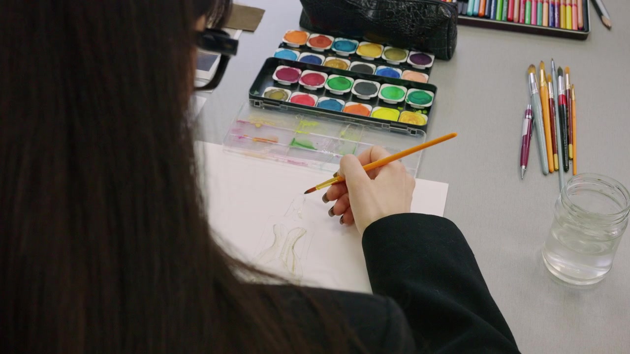 Close up of a hand painting a pale fashion figure sketch with an orange handled brush on white paper