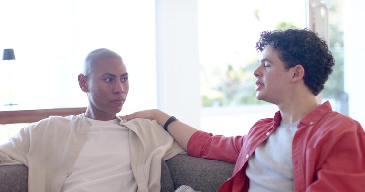 Diverse male friends on gray sofa, right man placing arm, telling story in red overshirt