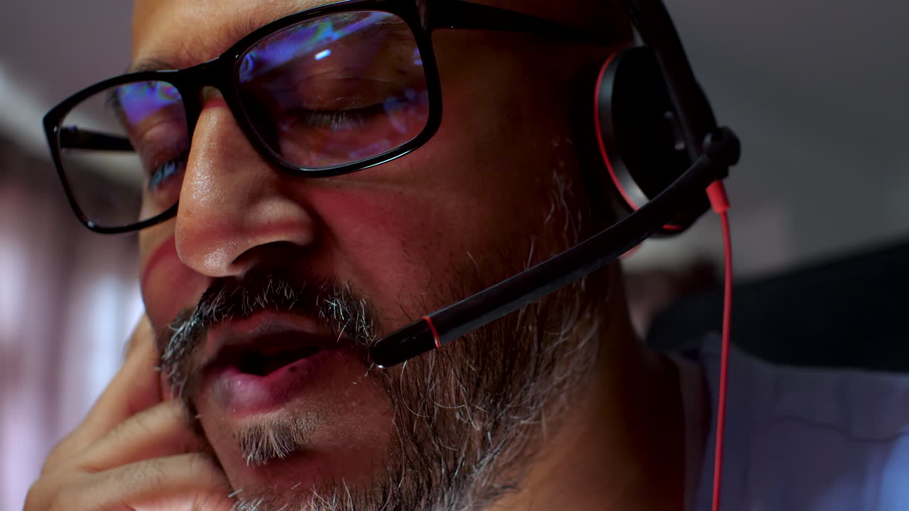 Close-up image of a South Asian man working, talking on a headset, and showing reflections in his glasses