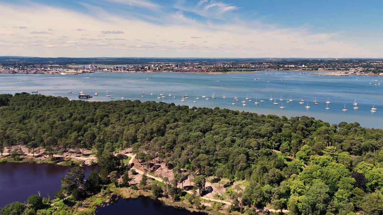 Flight over the lakes and woods of Brownsea Island Poole Harbour progressing to golden beach and clear water with views over the water with boats on moorings towards Poole itself on bright sunny day
