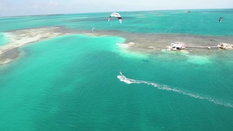 Vista-Aérea-Hombre-Kiteboard-Salto-Sobre-Agua-Azul-Clara-Frente-Palafito-Casa-Sobre-Pilotes,-Los-Roques