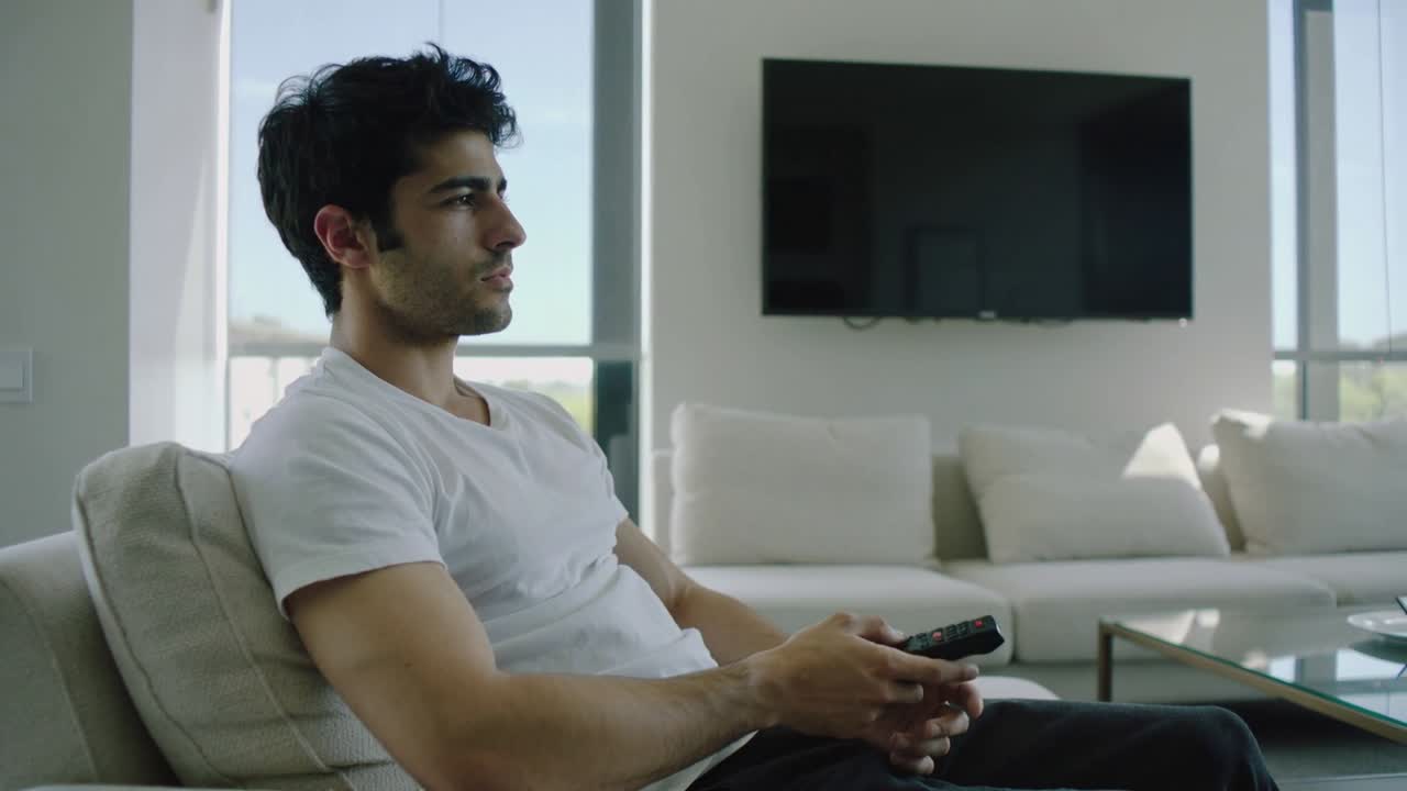 Uomo in maglietta bianca che guarda la TV, puntando il telecomando a casa per accenderla, con spazio per il testo.