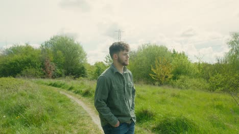 young man stands in open green space surrounded by low brush and overcast sky in natural scene, laughing with soft smile