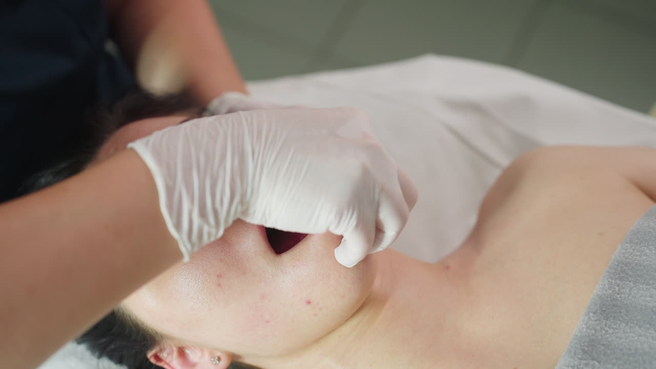 Close up of model under clinical blanket closing eyes in relaxed state as therapist wearing protective gloves dips fingertips into mouth and massages jawline with gentle care in clinic