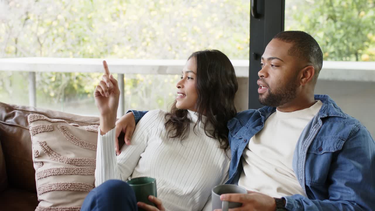 Diverse couple noticing window movement woman pointing at garden man listening with mugs on sofa