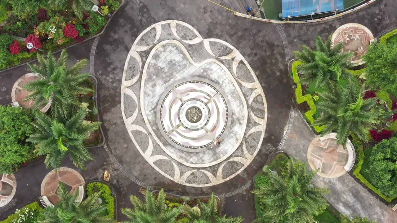 도시의 작은 공원에서 많은 열대 나무에 둘러싸인 가장자리에 앉아있는 두 사람과 함께 분수에 을 확대합니다.