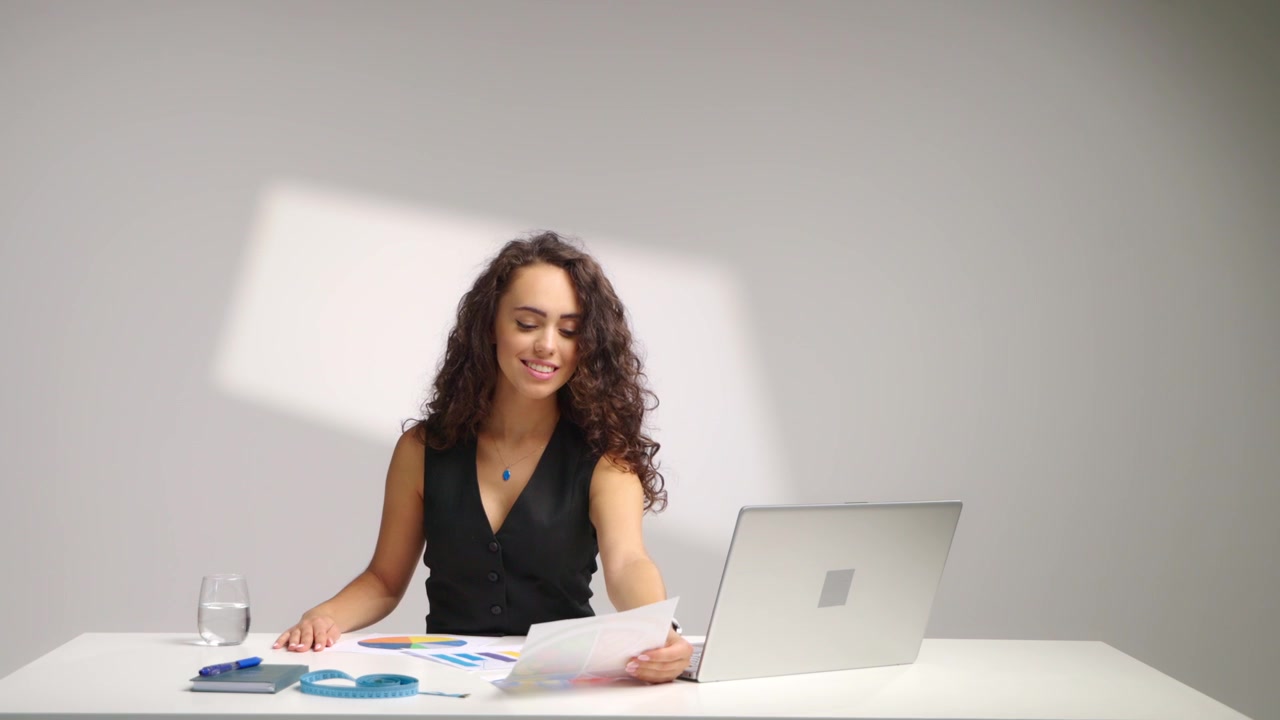Mujer sonriente sentada en un escritorio mirando la pantalla del portátil, levantando gráficos impresos y revisando documentos en la mesa. Consulta de nutrición, análisis de datos y proceso de planificación organizado en un estudio luminoso.
