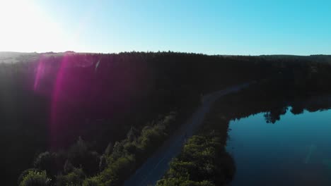 Aerial-shot-of-motorcycle-ride-on-a-road,-forest-and-lake-early-in-the-morning