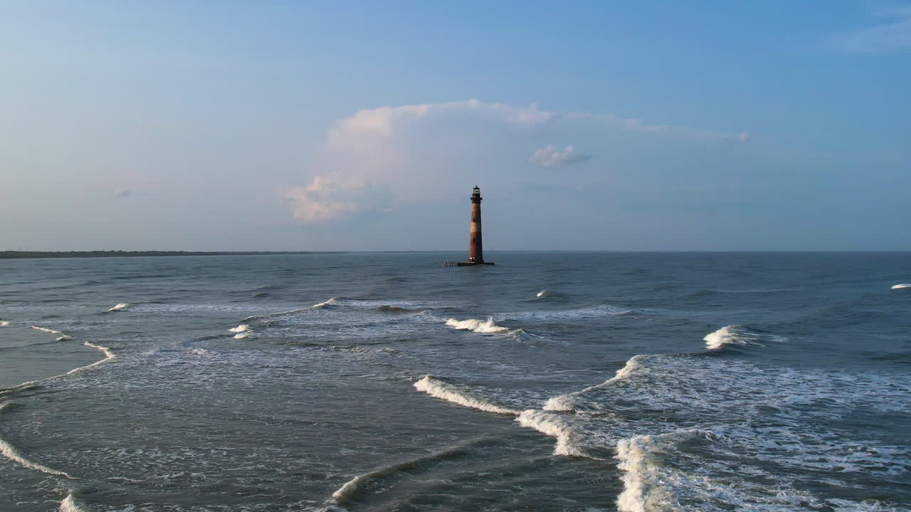 A drone shot moving towards a lighthouse that is surrounded by the ocean.