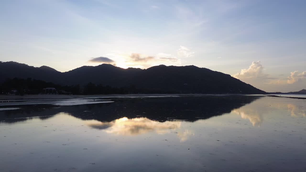 Drone footage capturing sunrise over Koh Phangan Thailand with the dark silhouette of a mountain and the reflection of sunlight shimmering on the calm tropical sea