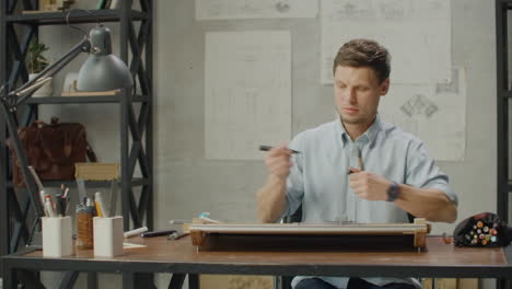 architect-man-Standing-working-with-blueprints-sketching-a-construction-project-on-wood-desk-at-home-office.Construction-design-concept.vintage-color-tone.-Engineer-works-in-a-bright-office-with-a-large-window-concentrates-and-draws-blueprints.-Workplace-of-an-architect-or-designer:-loft-style-minim