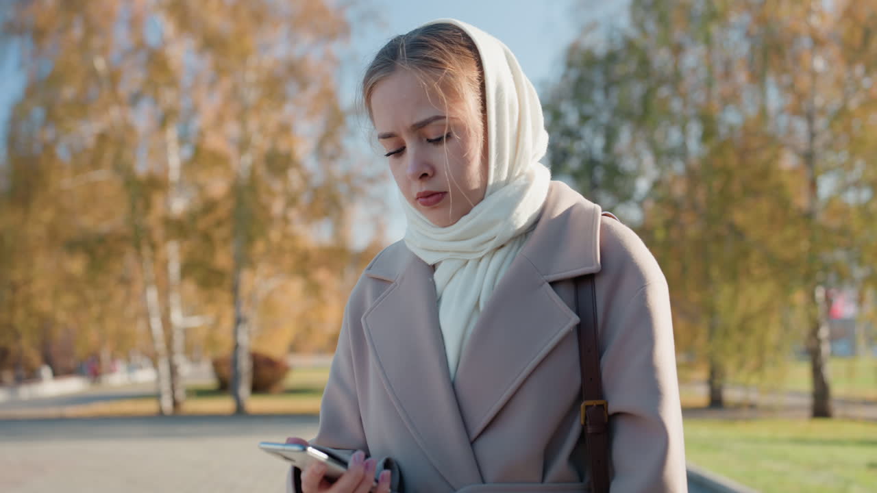 Hvid kvinde i efterårspark tjekker telefon, pakket ind i cremefarvet tørklæde og beige frakke, går langs fortovet nær birketræer og gyldne blade, fjerne bygninger skaber en rolig bystemning