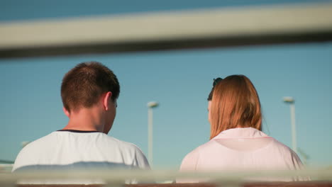 back view of lady with glasses on her head interacting with a man seated close on a bench, against a backdrop of iron railing, light pole, and bright sky