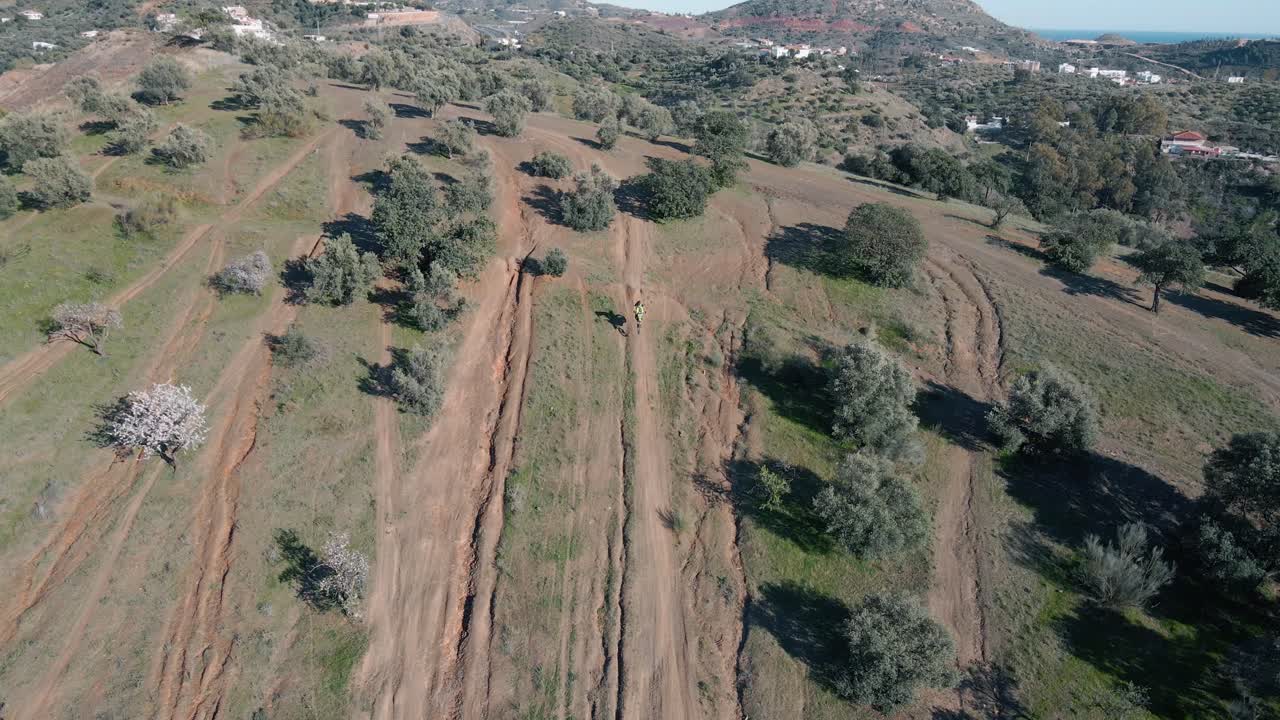 오토바이 운전자가 언덕 위에서 스턴트를 수행하는 동안 마른 먼지가 많은 트랙, 나무 및 산이 배경에 있는 말라가 스페인의 모터크로스 트랙에서 넓은 공중 정적 드론이 촬영되었습니다.