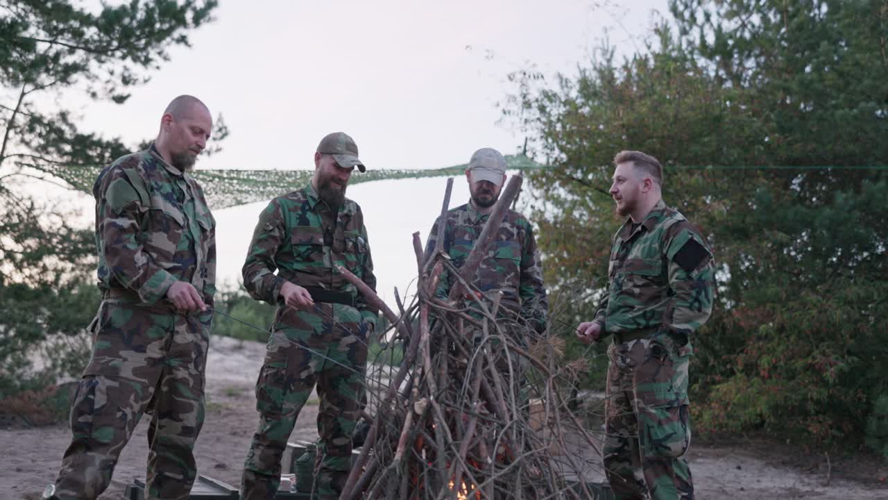 hombres militares con ropa de camuflaje y gorra se paran afuera alrededor de una fogata, hablando, contando historias, fritando salchichas en palos, descansando después del servicio, cazando en la base