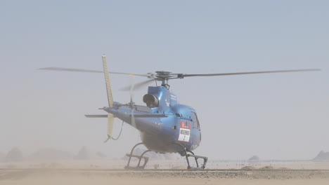 a blue airbus h125 helicopter taking off at the dakar rally with dusty landscape