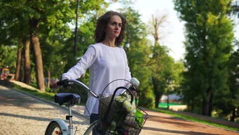 young woman in a white t-shirt and blue jeans walking on the cobblestone road in the city park holding her city bicycle