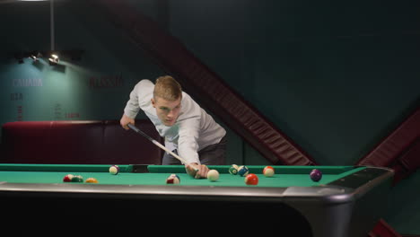 gamer in white shirt and grey trousers leans over green pool table, gripping cue stick with intense focus as he strikes cue balls. dark background enhances concentration