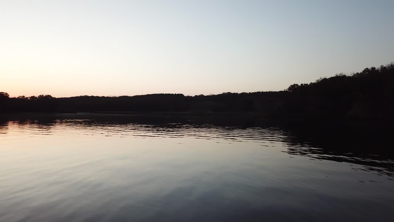 En fredfyldt skumringsstemning over Lake Wisconsin med blide krusninger, der reflekterer den blege himmel. Det stille vand strækker sig mod en mørk skovklædt kystlinje, hvilket fremkalder ro og naturlig skønhed.