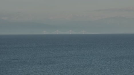 Long-range-telephoto-shot-of-a-far-away-bridge-with-mountains-in-the-background-and-sea-in-the-foreground