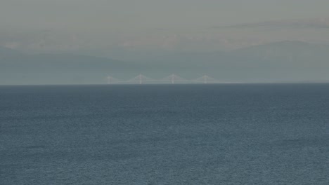 Toma-De-Teleobjetivo-De-Largo-Alcance-De-Un-Puente-Lejano-Con-Montañas-En-El-Fondo-Y-El-Mar-En-Primer-Plano