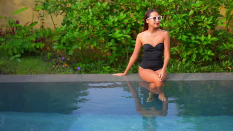 Sexy-young-healthy-woman-sitting-on-the-edge-of-a-private-swimming-pool-on-lush-greenery-background,-daytime-slow-motion-handheld