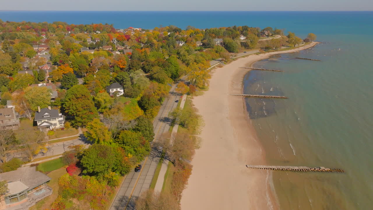 Drone aerial pulling back over a car driving along the shoreline road in Sheboygan, Wisconsin, with neighborhood houses and autumn trees beside the beach, piers, and Lake Michigan
