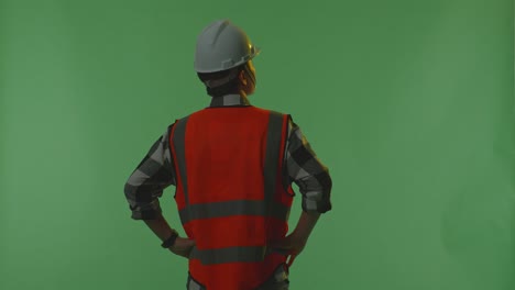 back view of a male engineer wearing safety helmet looking around while standing with arms akimbo in the green screen background studio, during sunset or sunrise time